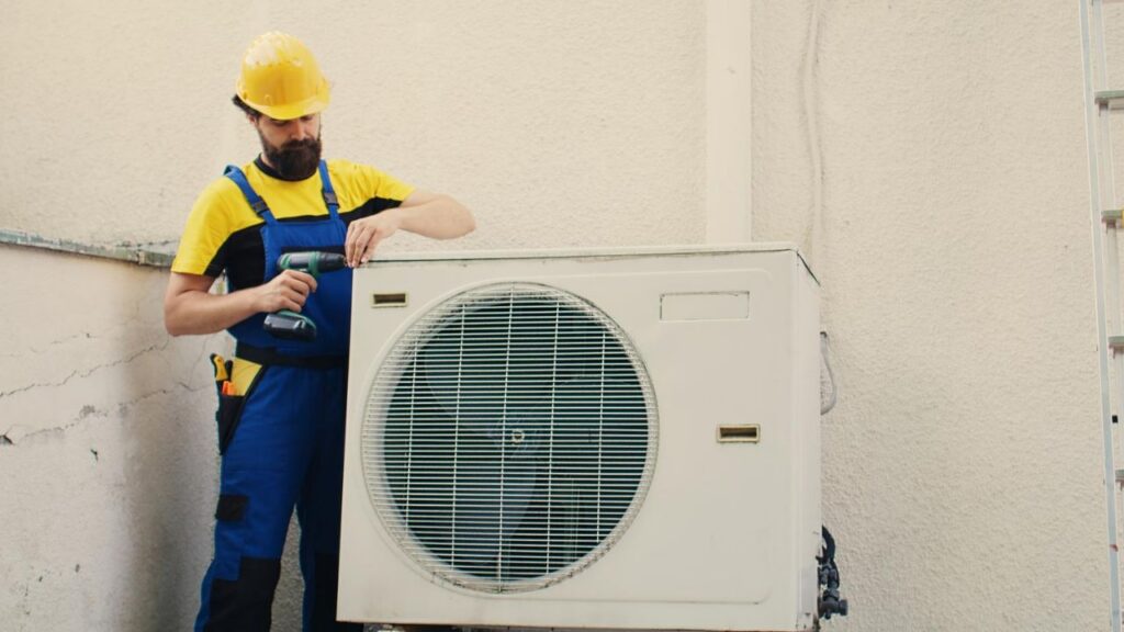 HVAC technician using a cordless drill while servicing an outdoor air-conditioning condenser unit.