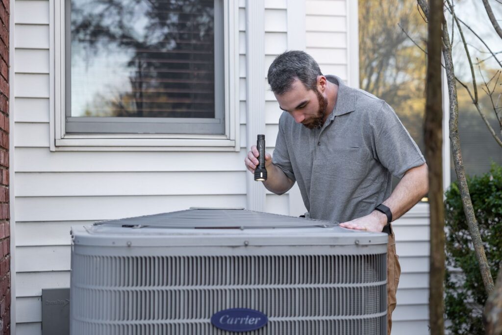 HVAC technician inspecting an outdoor air-conditioning condenser unit with a flashlight.